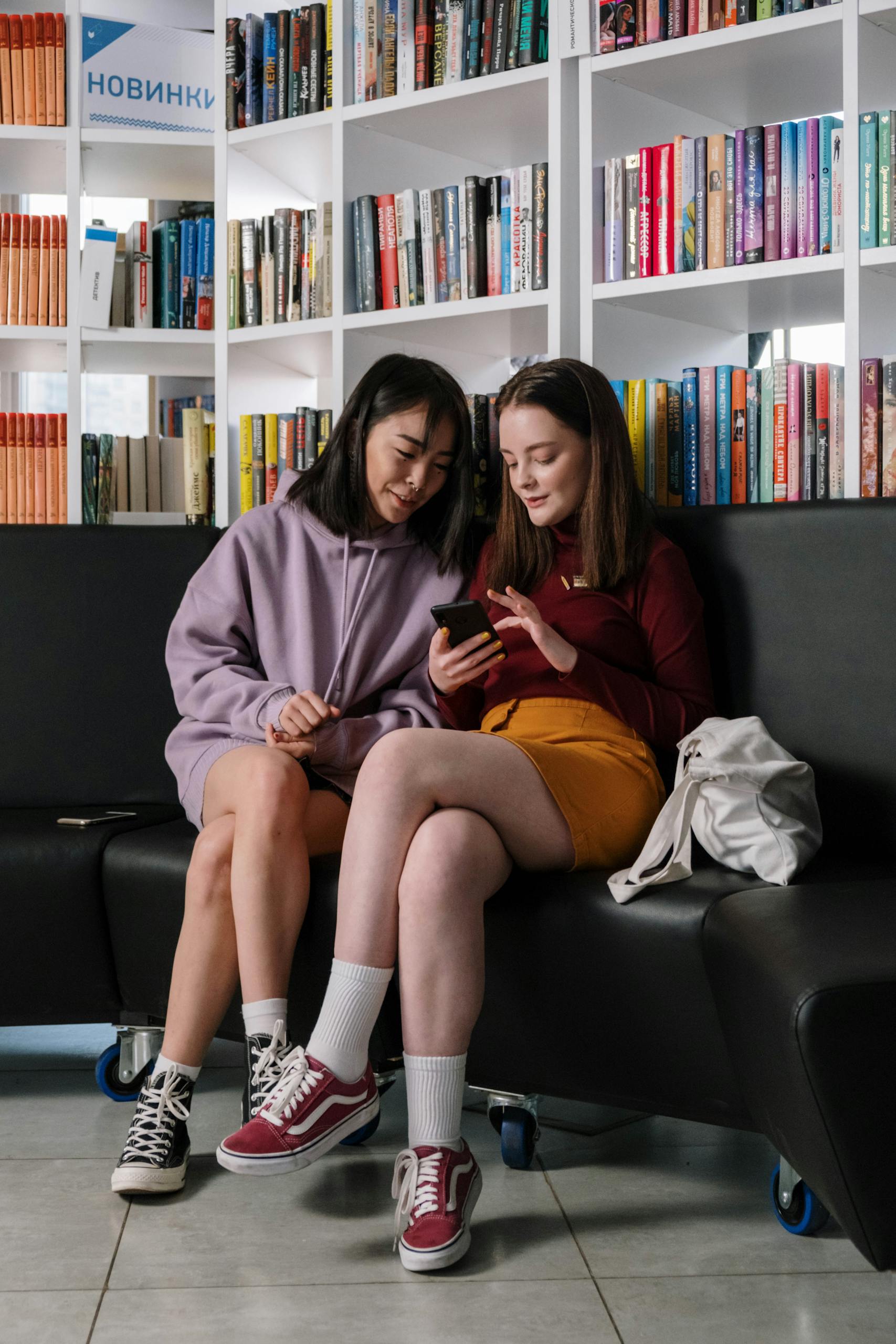 Two teenage girls sitting and talking in a library, engaging with a smartphone.