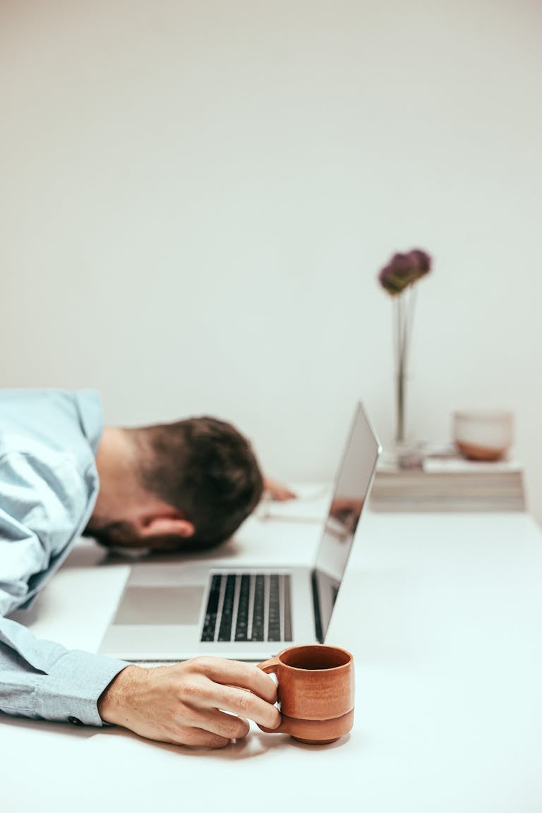A fatigued man in an office setting asleep at his desk with a laptop and coffee mug.