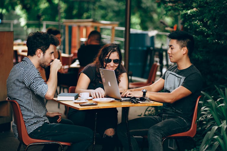 A group of young adults working on a laptop at an outdoor coffee shop, enjoying teamwork and collaboration.